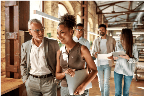 A diverse group of professionals walking in a modern office, discussing work, holding laptops and documents, in a collaborative, industrial-style setting.
