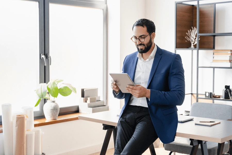 A man in a blue blazer sits on a desk, looking at a tablet in an office enviroment.