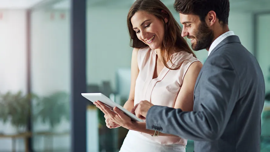 A businesswoman and businessman smiling and working on a computer tablet in an office environment.