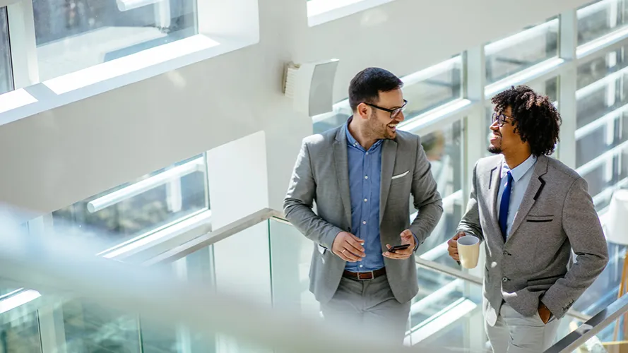 Two young businessmen ascending a staircase and talking in an office setting.