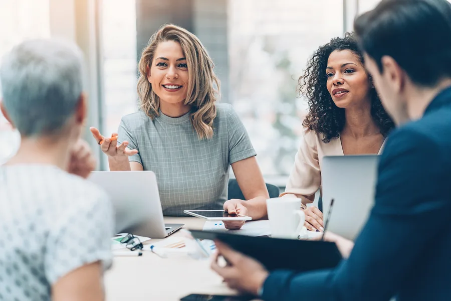 A diverse group of professionals in a meeting, with a smiling blonde woman speaking and holding a tablet. Laptops, documents, and a coffee cup are on the table in a bright, modern office.