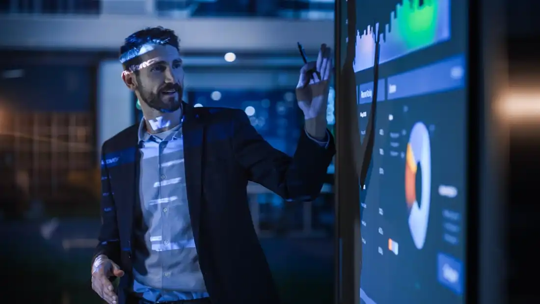 Man in a suit pointing to data charts and graphs on a large screen during a presentation.
