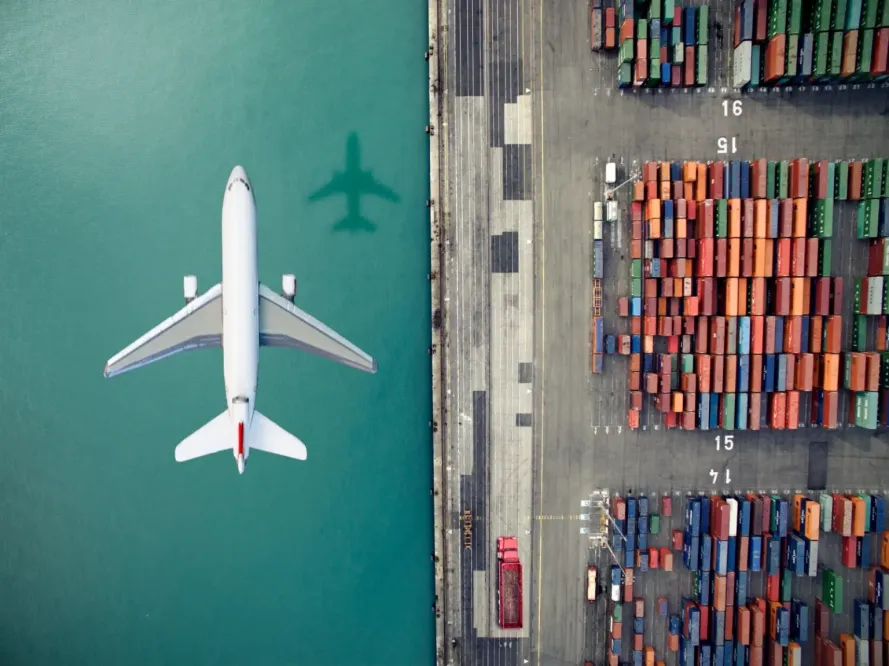An overhead view of a jet plane flying over a waterway with colorful shipping containers stacked nearby.